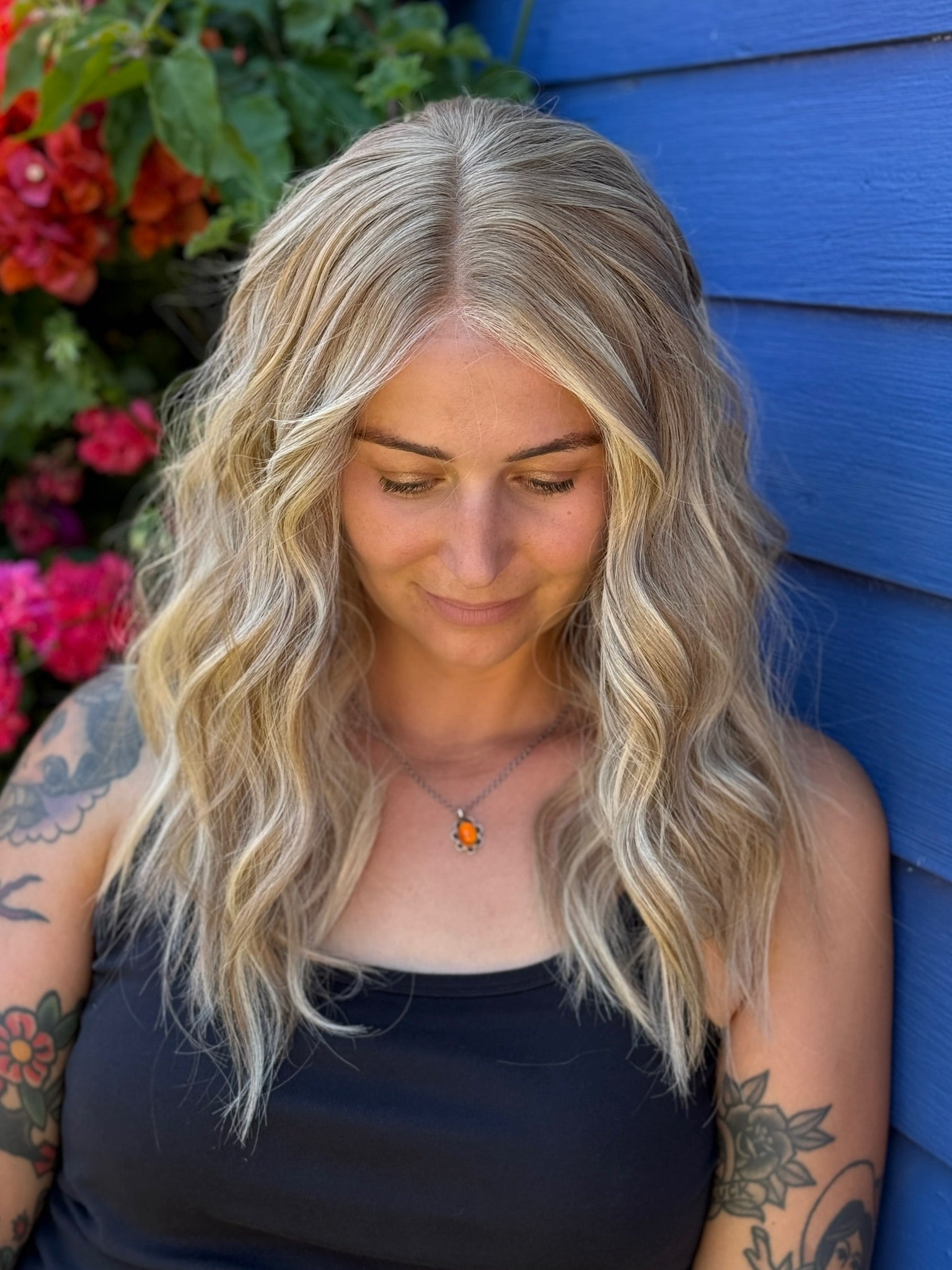 Woman with long blonde wig and tattoos standing against a blue wall with flowers in the background