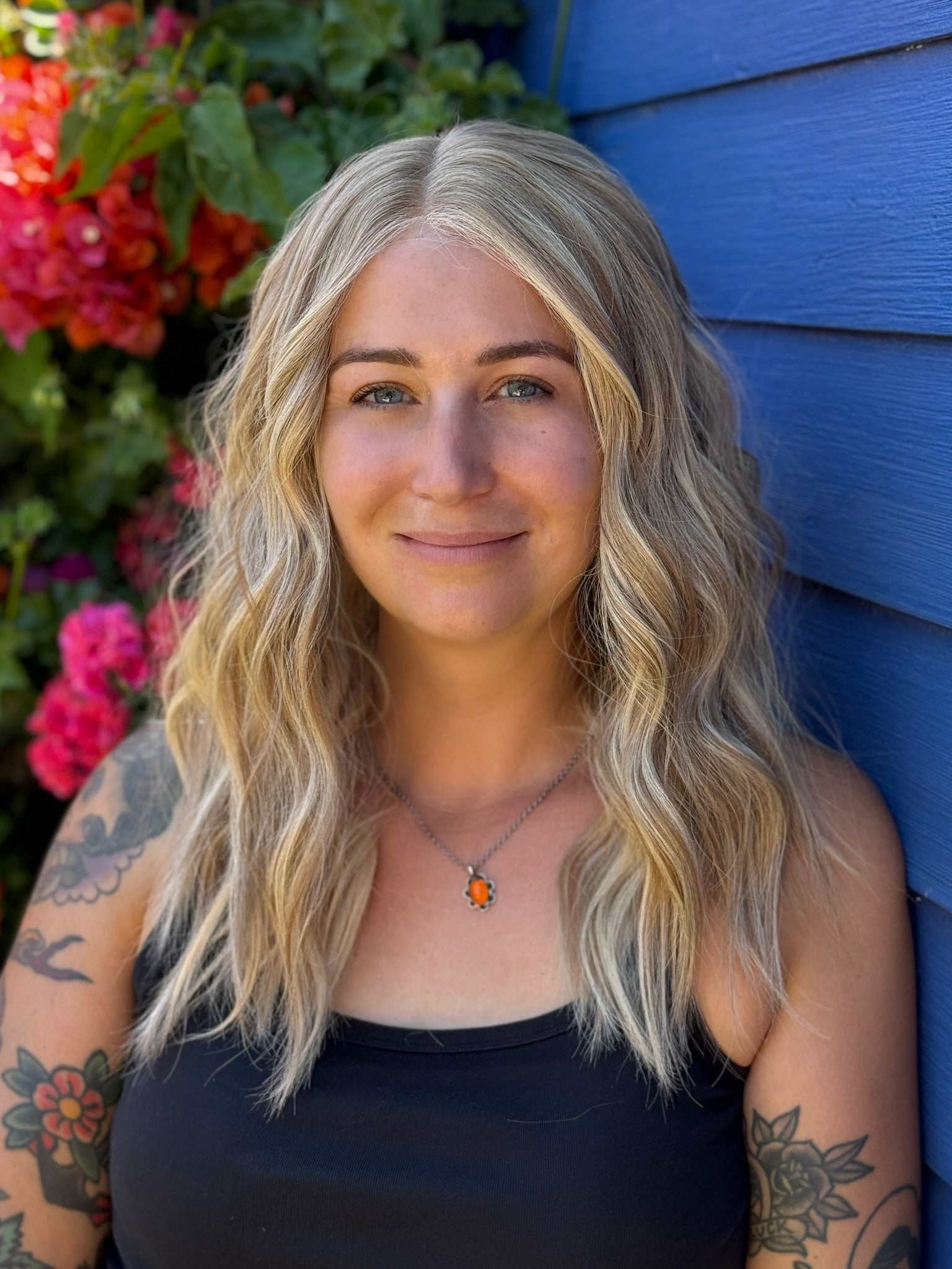 Woman with blonde wig and tattoos standing against a blue wall with flowers in the background